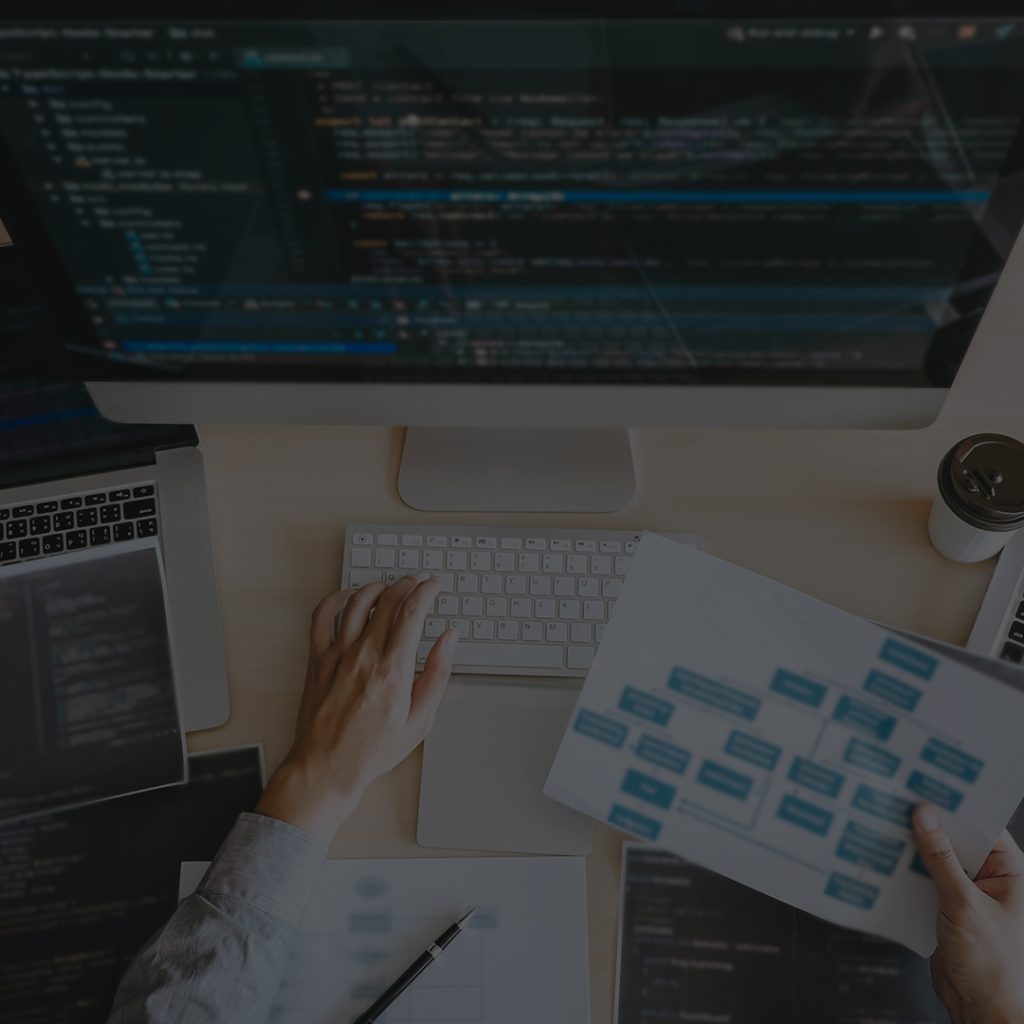 Developer workspace: hands on a white keyboard in front of a large monitor displaying code, with a laptop, papers, and coffee cup nearby.