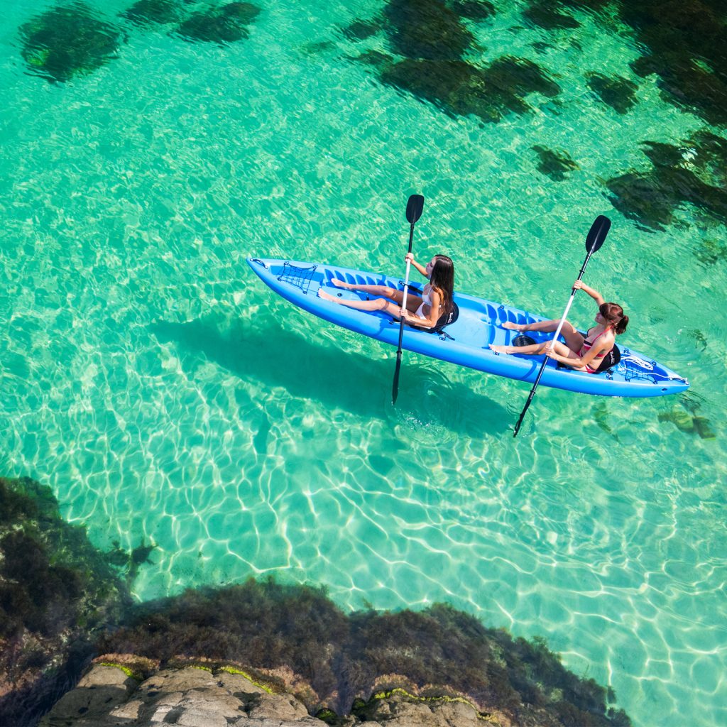 Two people paddling a blue tandem kayak in clear turquoise water near a rocky shore.